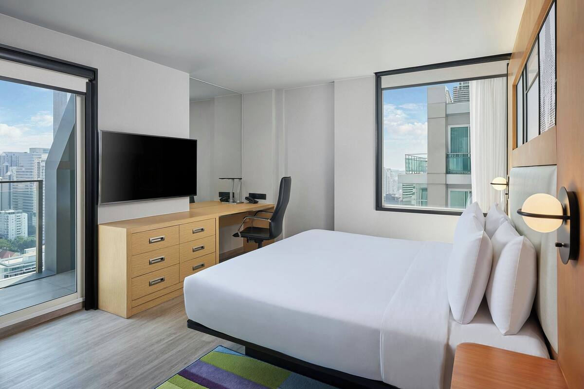 A modern hotel room with a large bed, a desk with a flat-screen TV, a chair, and a view of the city through large windows.