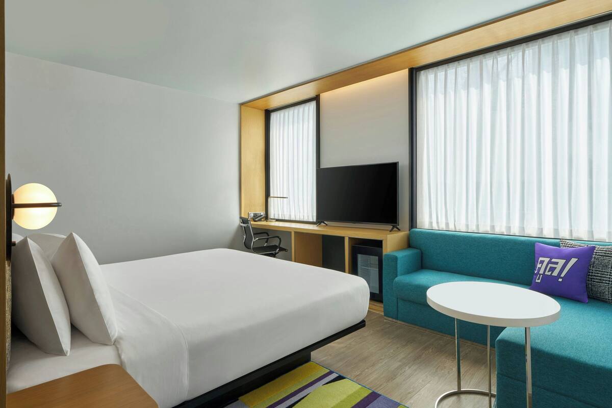 A hotel room with a bed, a sofa, a TV, a small table, and a window with curtains.