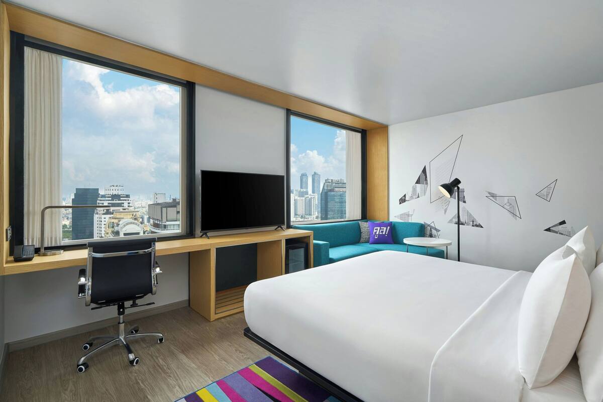 A hotel room with a large bed, a desk with a computer, a TV, and a view of the city skyline.