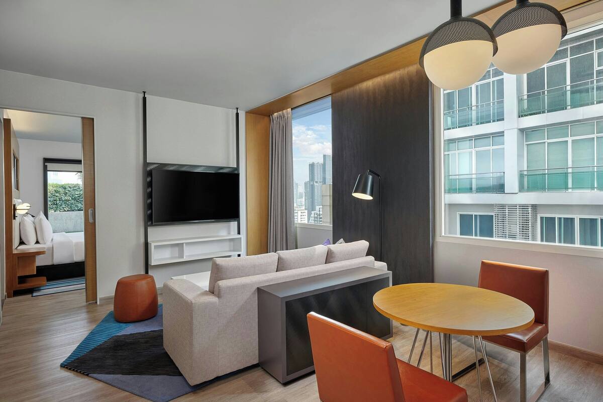 A modern hotel room with a sofa, a dining table, a TV, and a large window with a city view.