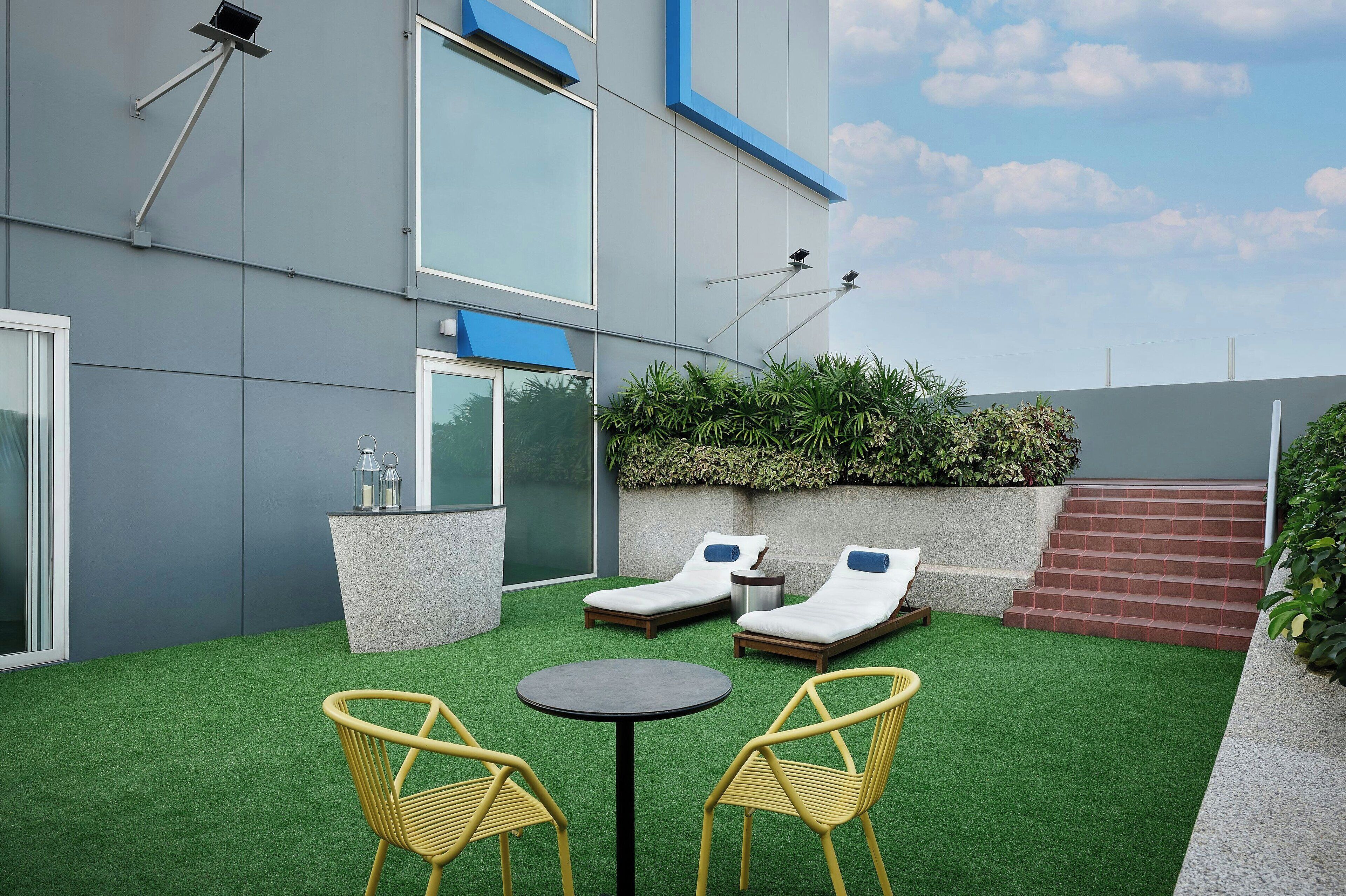 Un patio exterior moderno con césped artificial, dos sillones, una mesita redonda y una jardinera de concreto con botellas.