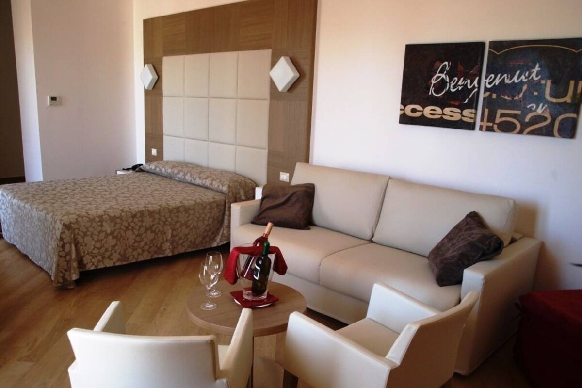 A hotel room with a bed, a sofa, a small table with a bottle and glasses, and a wall with a sign.