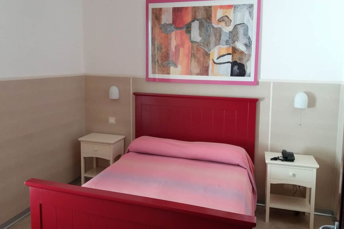 A room with a red bed, a small bedside table, a nightstand with a phone, and a painting on the wall.
