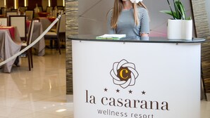 Mostrador de recepción en La Casarana Wellness Resort, con un logotipo, una orquídea en maceta y una vista parcial del comedor interior.