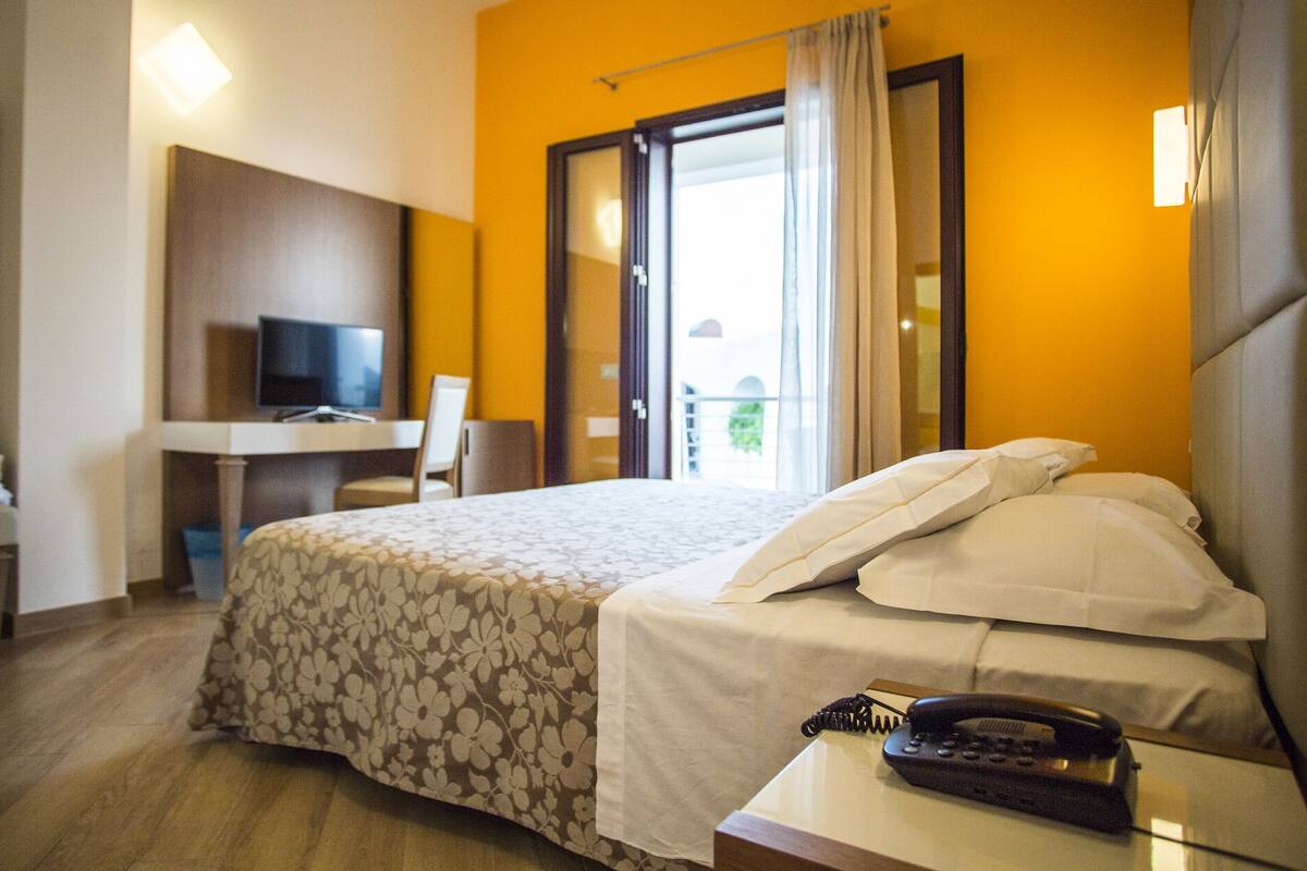 A hotel room with a bed, a desk with a TV, and a balcony with curtains.