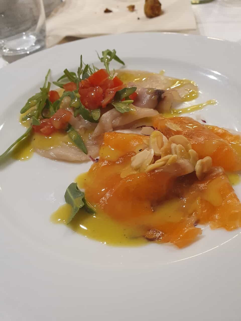 Ein weißer Teller mit einem Fischgericht, möglicherweise Lachs, garniert mit Kräutern und Nüssen, dazu gewürfelte Tomaten.
