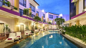 Un área de piscina hotelera con sillones de descanso, una piscina y varios balcones con plantas.