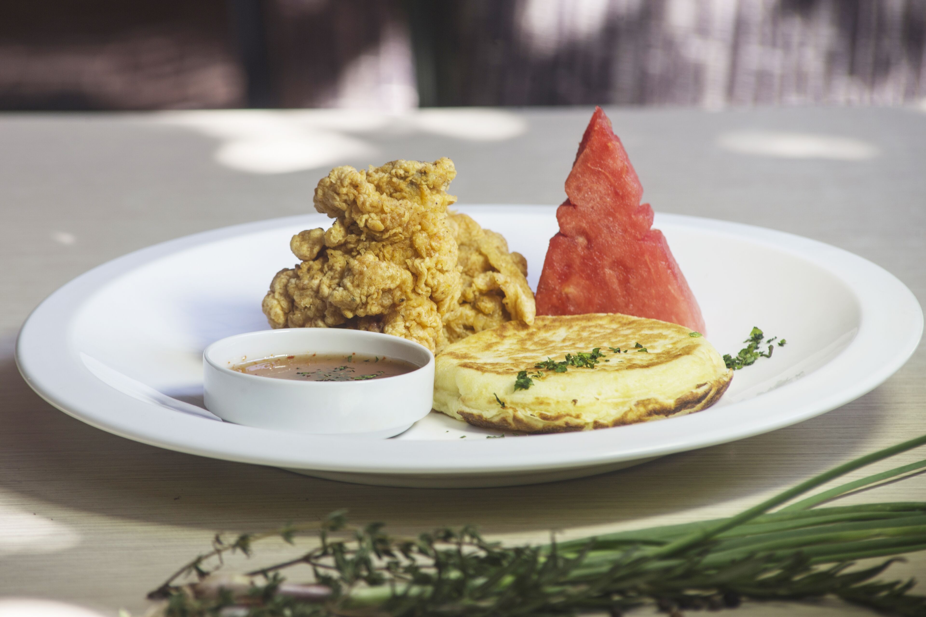 A plate with a piece of fried chicken, a slice of watermelon, and a small bowl of sauce on a wooden table.