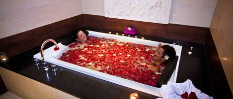 A bathtub filled with rose petals and candles, with a towel and a pink gift box on the side.