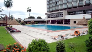 A swimming pool with lounge chairs and umbrellas, surrounded by greenery and a building with multiple floors and balconies. - Presidential Hotel (Port Harcourt)