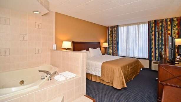 Une chambre d’hôtel dotée d’une grande baignoire, d’un lit avec une tête de lit en bois, de deux lampes de chevet, d’un bureau avec un ordinateur et d’une fenêtre avec des rideaux.
