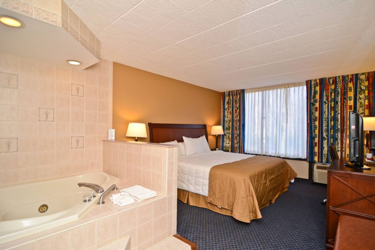 A hotel room with a large bathtub, a bed with a wooden headboard, two bedside lamps, a desk with a computer, and a window with curtains.