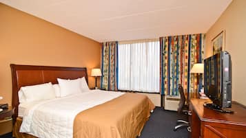 Quarto de hotel com uma cama grande, uma mesa com computador e uma janela com cortinas.
