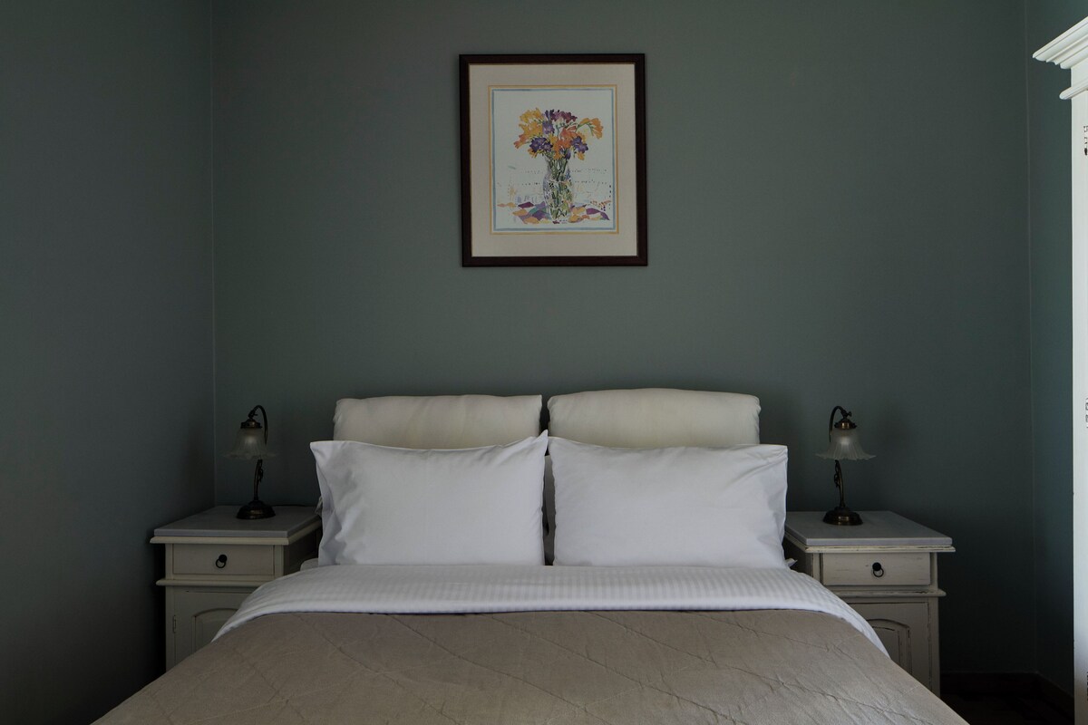 A bedroom with a bed, two bedside tables with lamps, a framed picture on the wall, and a nightstand with a drawer.
