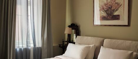 Una cama bien hecha con sábanas blancas, una mesita de noche con una lámpara y un cuadro floral enmarcado en la pared.