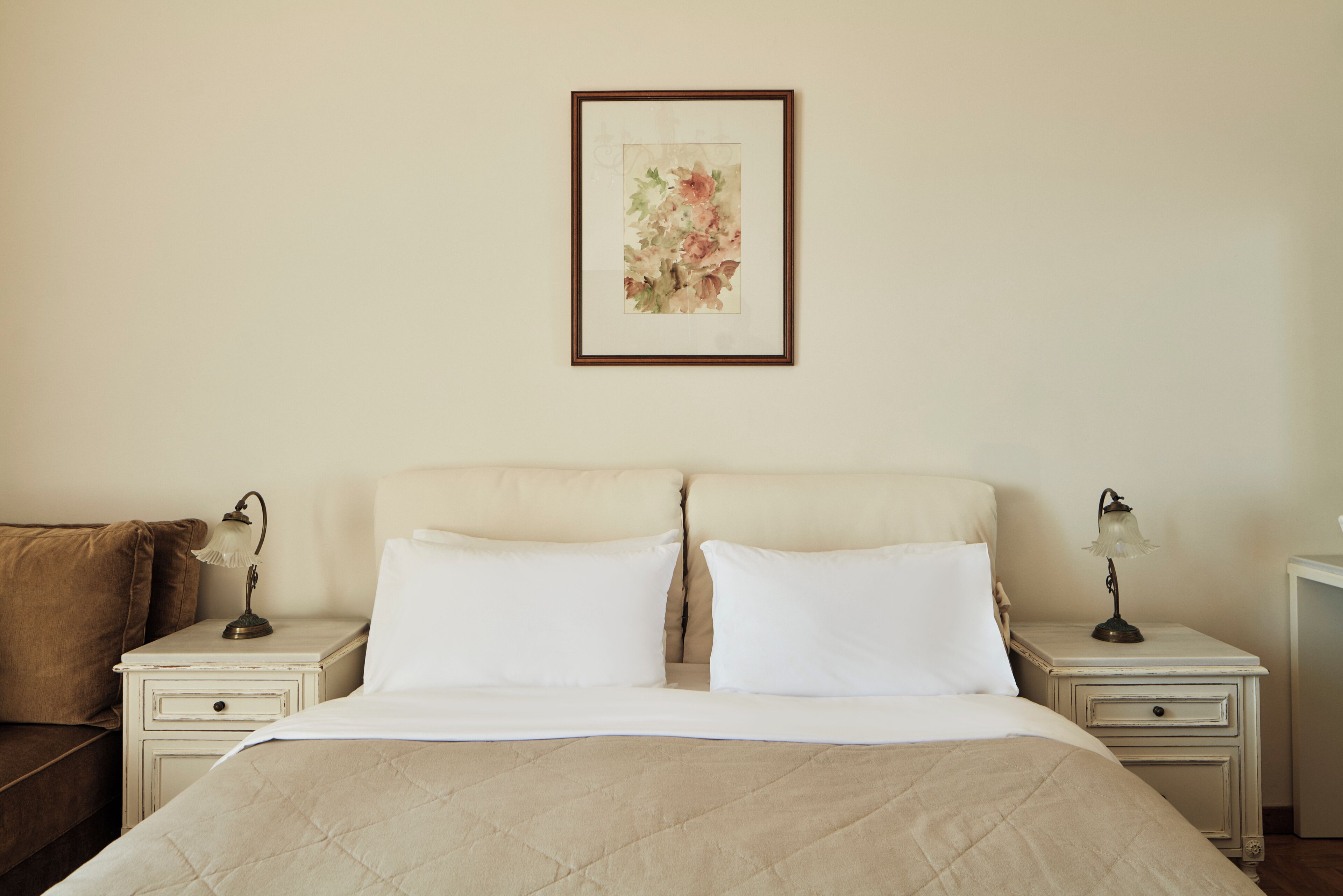 Una cama bien hecha con cubrecama acolchada, dos mesitas de noche con lámparas, una obra de arte floral enmarcada en la pared y un sof á marrón al fondo.