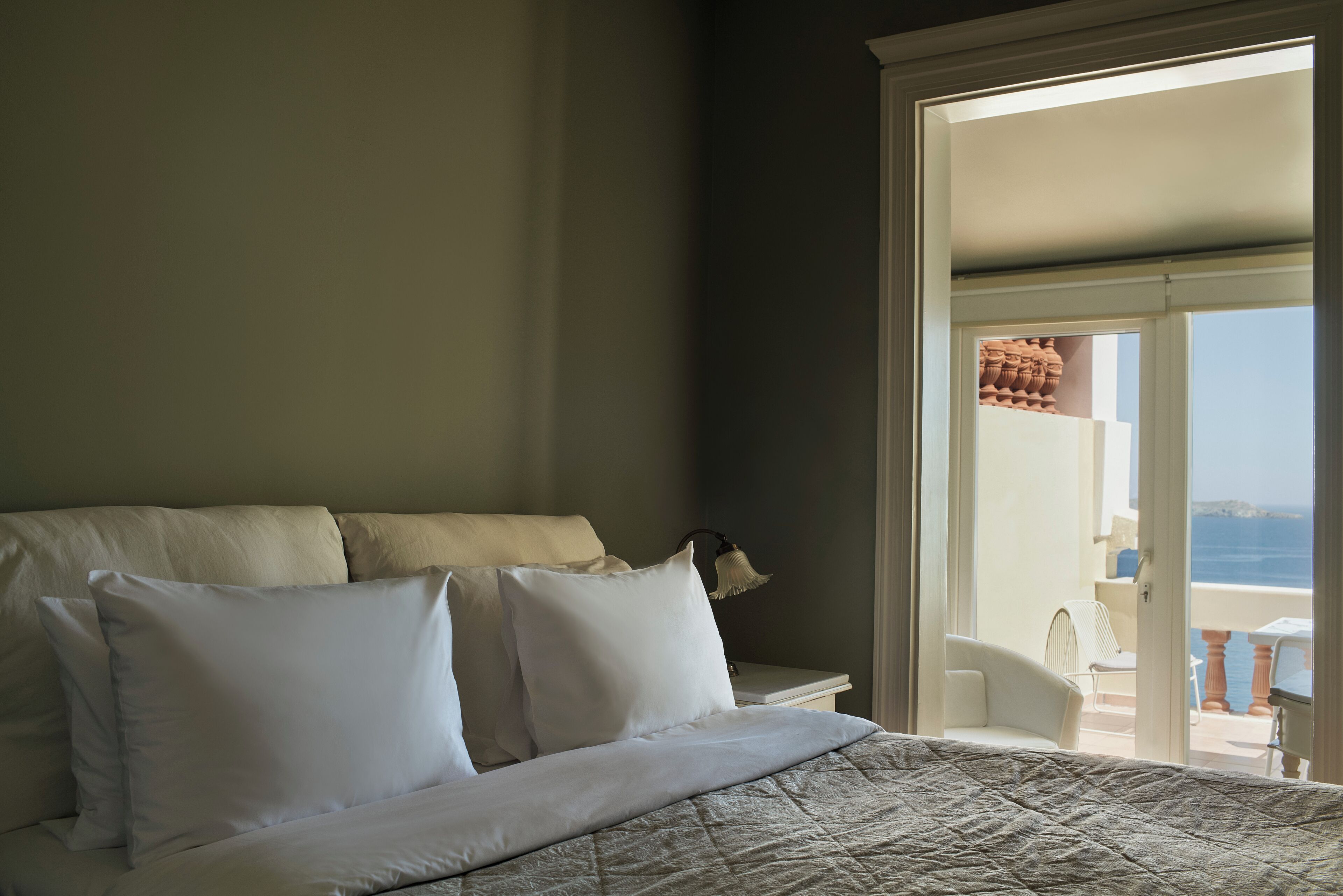 Quarto com uma cama grande, roupa de cama branca e vista para o mar através de uma porta aberta.