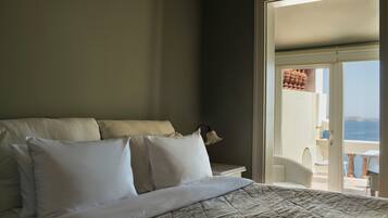Quarto com uma cama grande, roupa de cama branca e vista para o mar através de uma porta aberta.