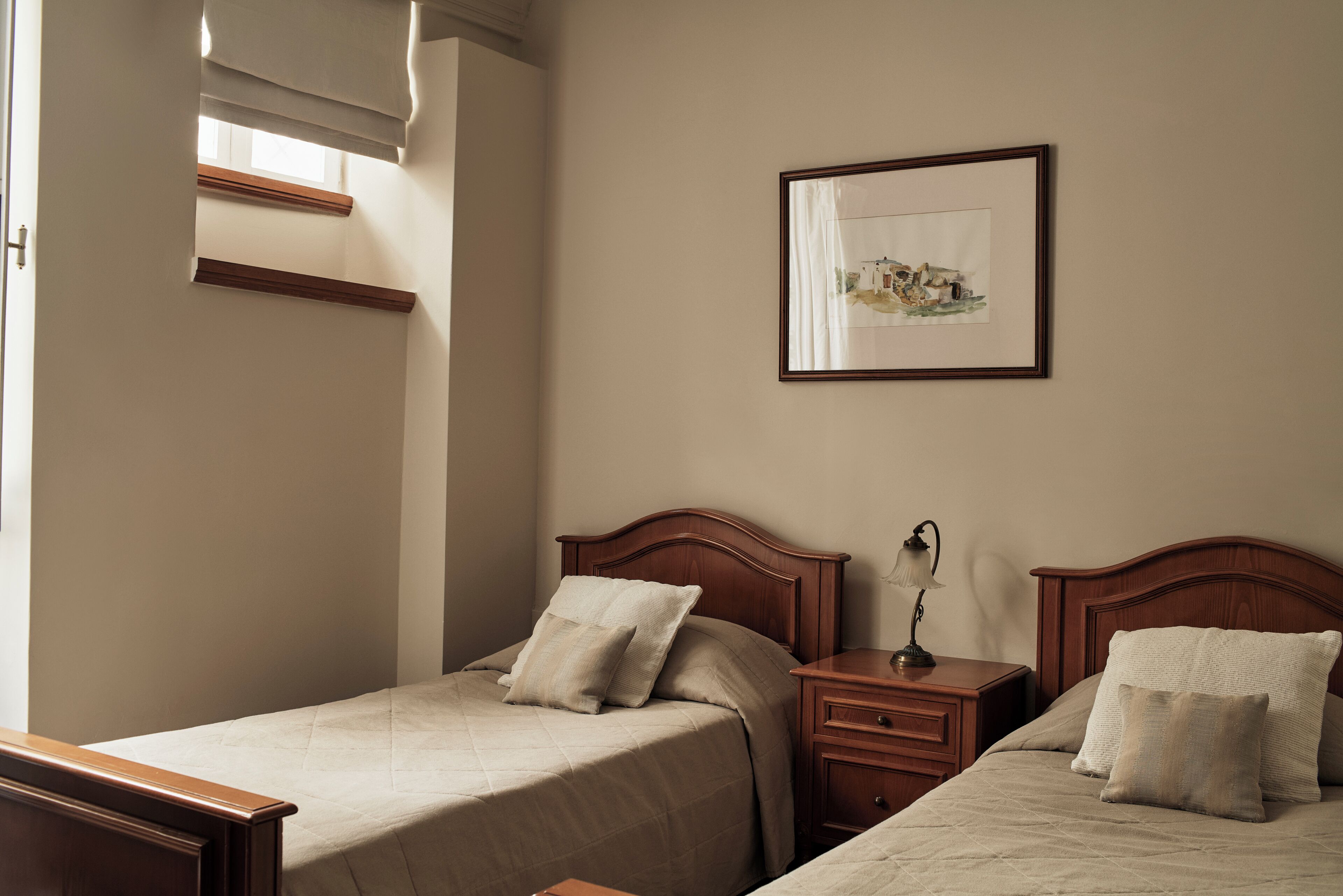 A bedroom with two beds, a nightstand with a lamp, a framed picture on the wall, and a window with blinds.