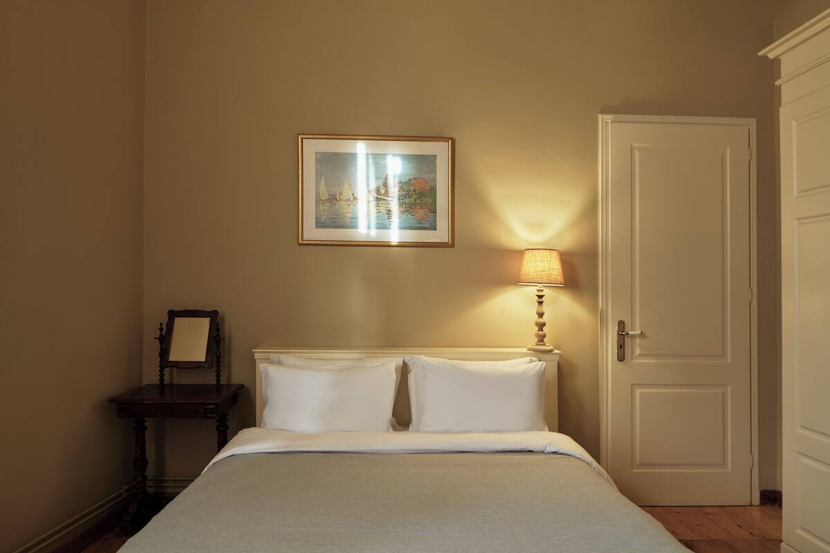 Una cama bien tendida con sábanas blancas, una mesita de noche con lámpara, un cuadro en la pared y una puerta cerrada.