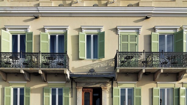 A two-story building with a symmetrical facade, featuring a central entrance with a wooden door, green shutters, and a balcony with wrought iron railings.