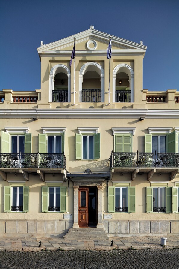 A two-story building with a symmetrical facade, featuring a central entrance with a wooden door, green shutters, and a balcony with wrought iron railings.