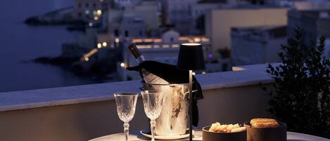 A table with two glasses, a bottle of champagne, a bowl of snacks, and a candle on a balcony overlooking a marina.