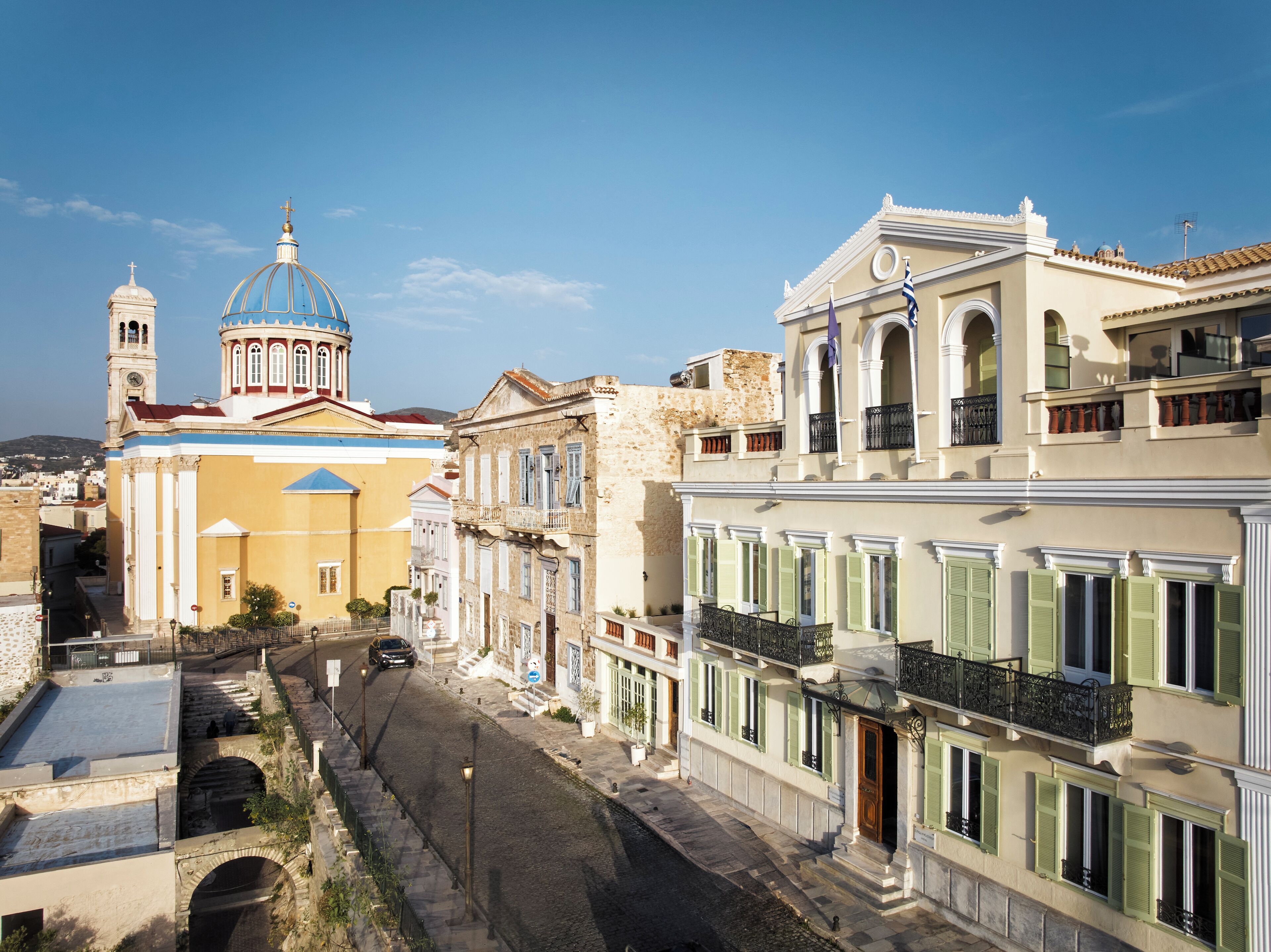 A street with historic buildings, including a church with a dome and bell tower, and a row of multi-story buildings with balconies.