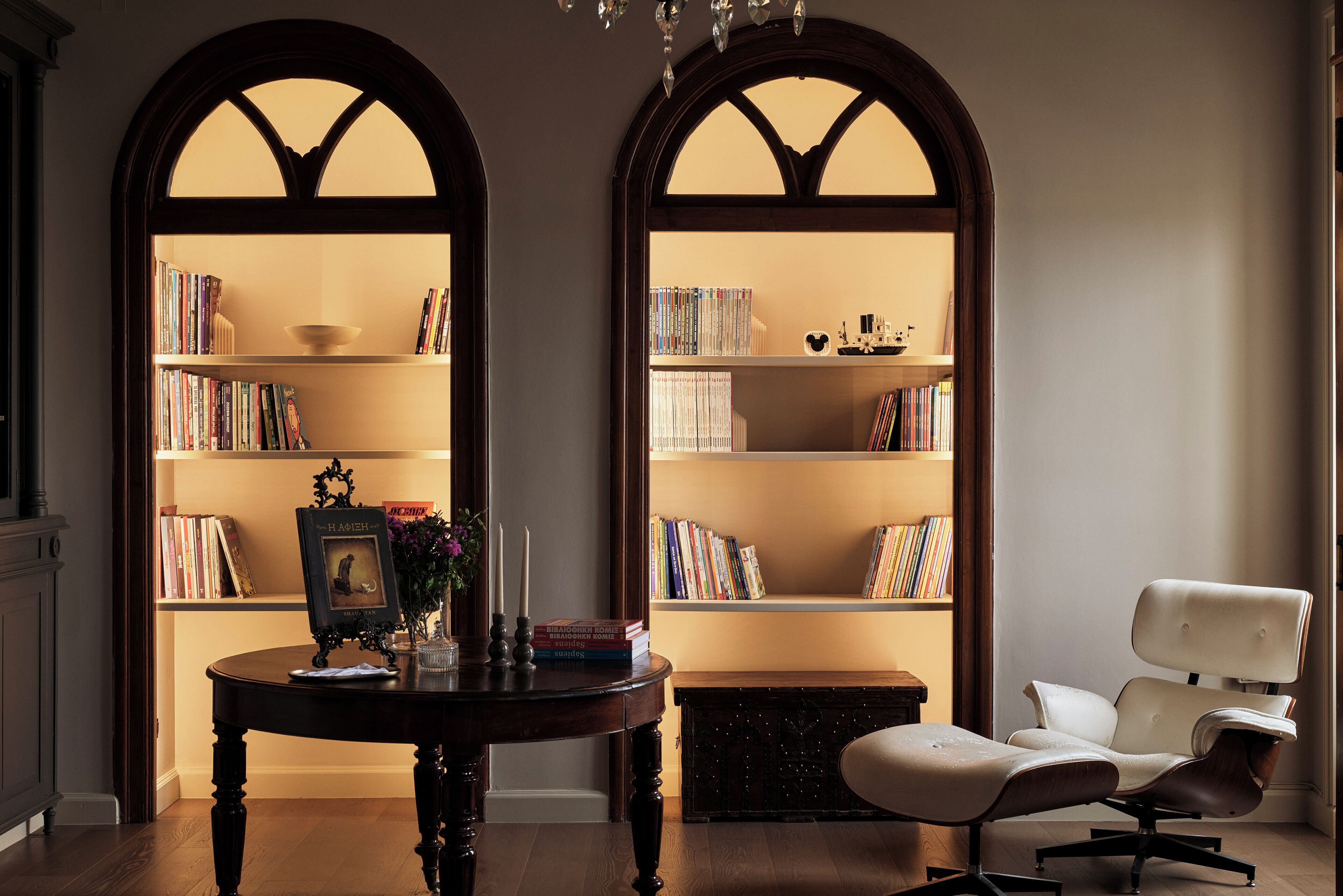 A library with two tall bookshelves, a round wooden table with books and a framed picture, a white armchair, and a dark wooden chest of drawers.