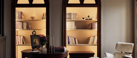 A library with two tall bookshelves, a round wooden table with books and a framed picture, a white armchair, and a dark wooden chest of drawers.