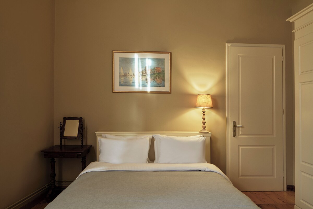 A bedroom with a bed, a bedside table with a lamp, a framed picture on the wall, and a door.