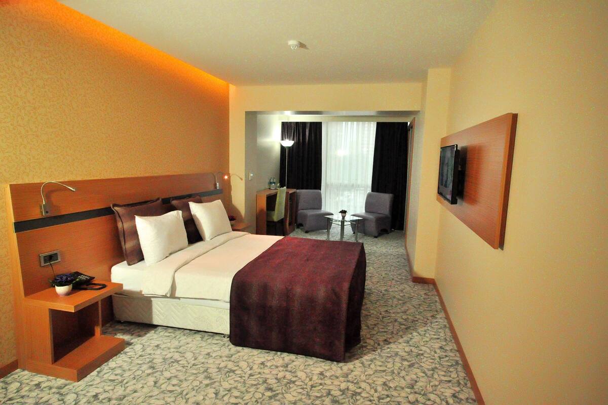 Hotelkamer met een bed, nachtkastje, lamp, televisie en een zithoekje met stoelen en een tafel.