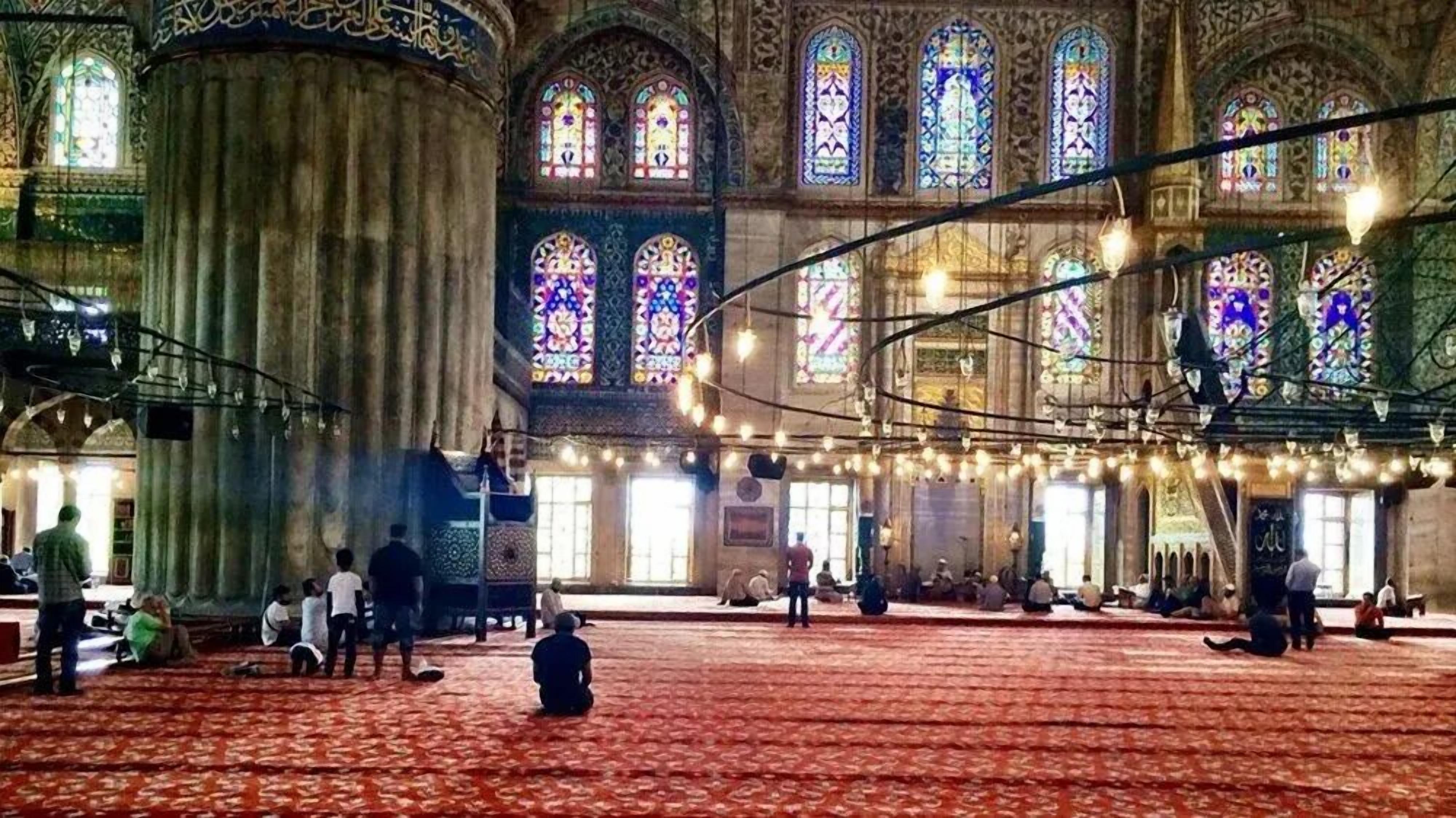 An interior of a historic building with large stained glass windows, a patterned carpet, and a high ceiling.