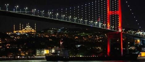 Una vista nocturna de un puente con una torre iluminada en rojo, una mezquita con minaretes iluminados y un paisaje urbano al fondo.