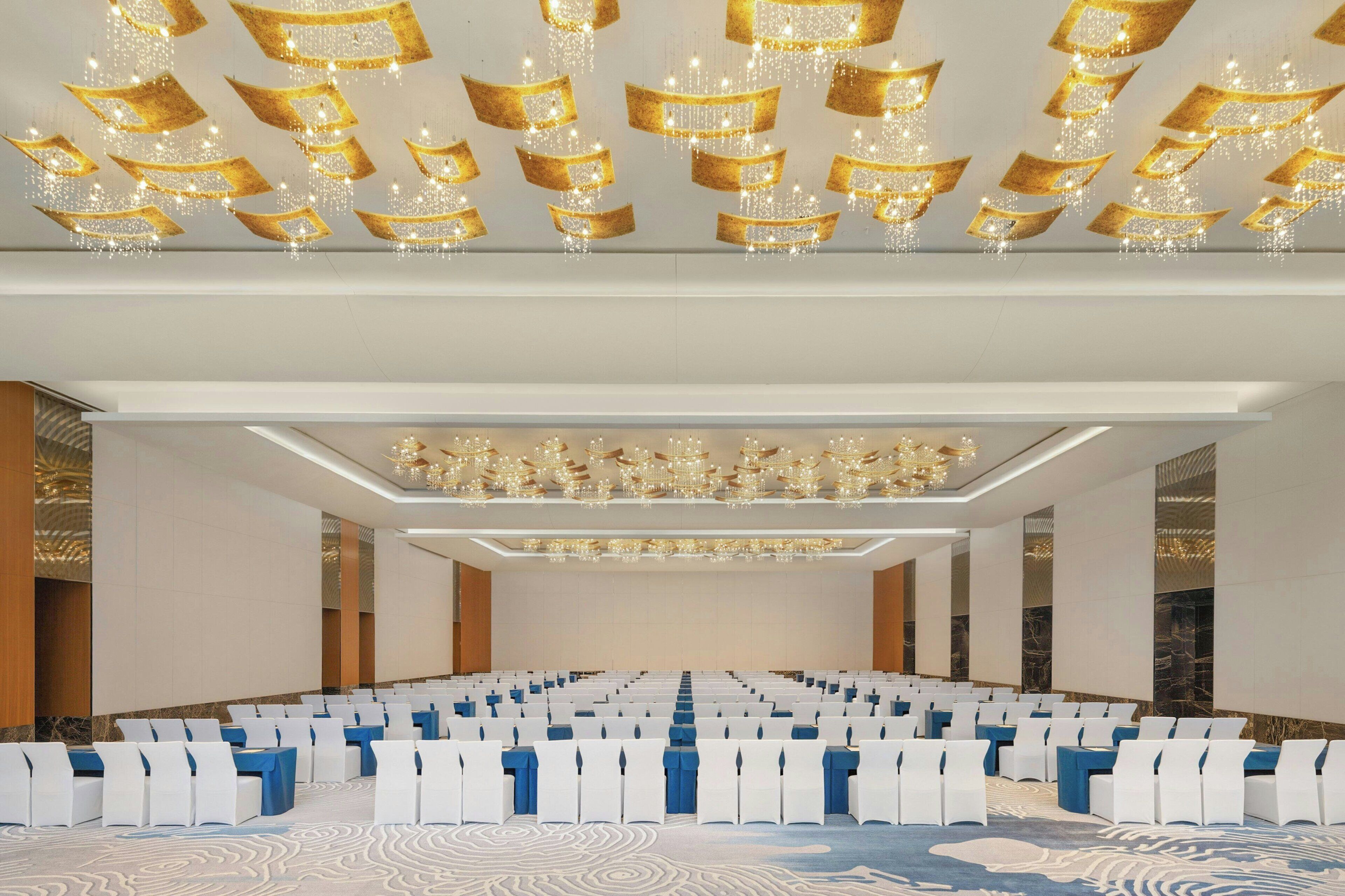 Une salle de conférence avec des rangées de chaises blanches, des nappes bleues et des lustres décoratifs.
