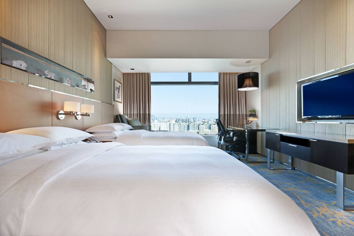 A hotel room with two beds, a desk, a TV, and a city view.