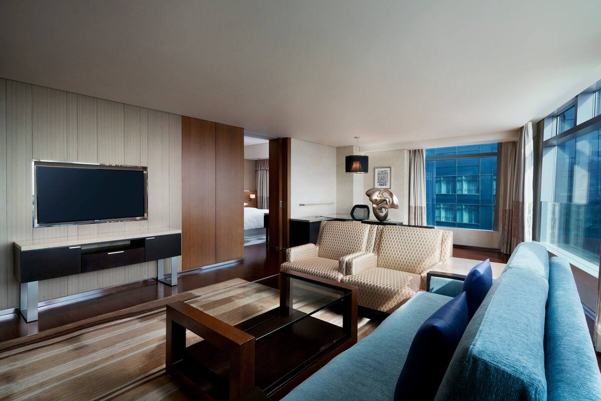 A modern hotel room with a large flat-screen TV mounted on the wall, a sofa, a patterned armchair, and a coffee table.