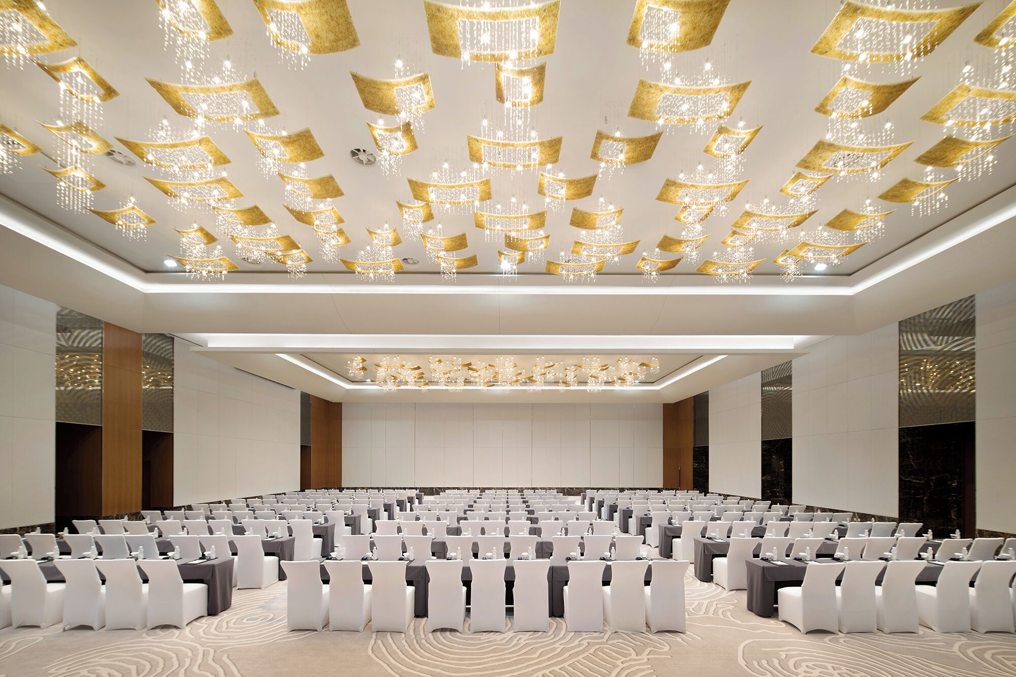 Une salle de conférence avec des rangées de tables et de chaises, une moquette à motifs et un plafond orné de lustres suspendus.