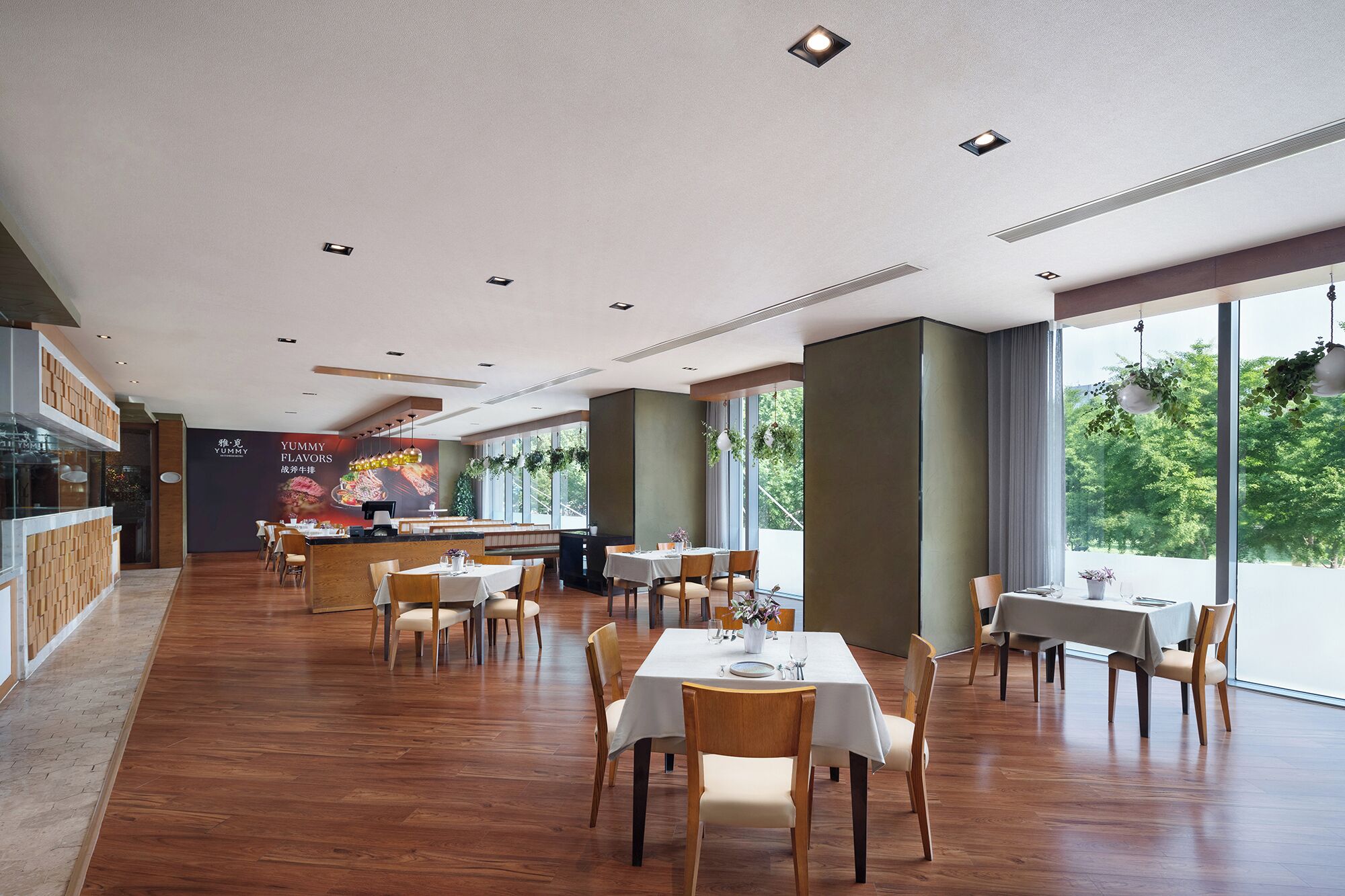 A restaurant interior with wooden flooring, tables with white tablecloths, and a large window offering a view of greenery.