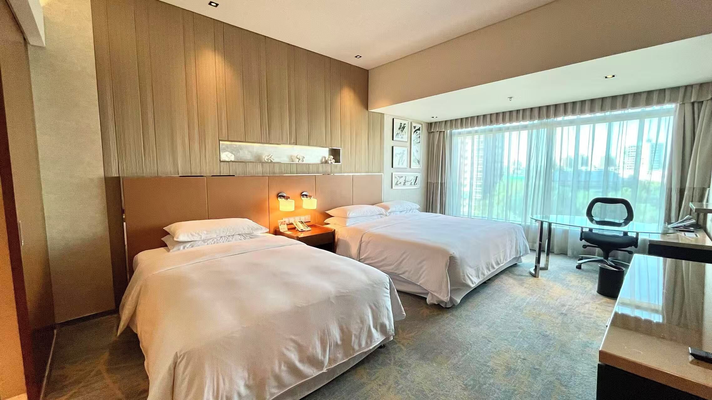 Ein Hotelzimmer mit zwei Betten, einem Schreibtisch, einem Stuhl und einem großen Fenster mit Vorhängen.