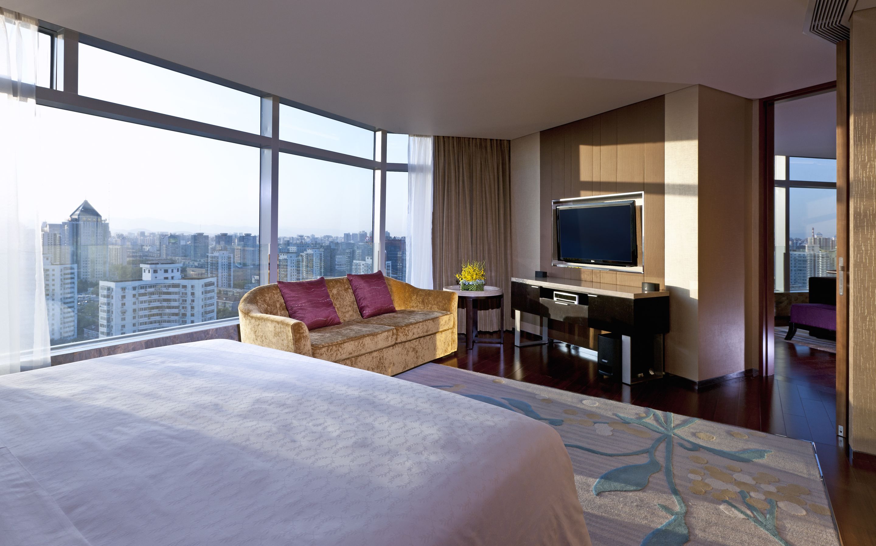 A hotel room with a large bed, a sofa, a desk with a TV, and a city view.