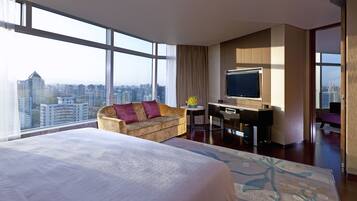 A hotel room with a large bed, a sofa, a desk with a TV, and a city view.