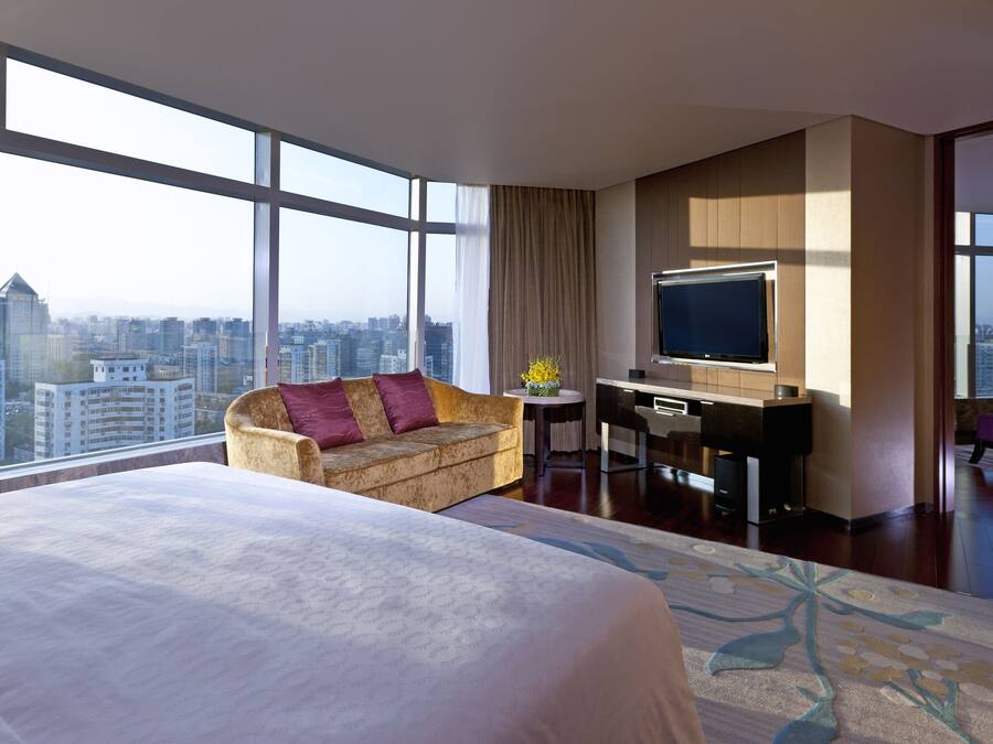A hotel room with a large bed, a sofa, a desk with a TV, and a city view.