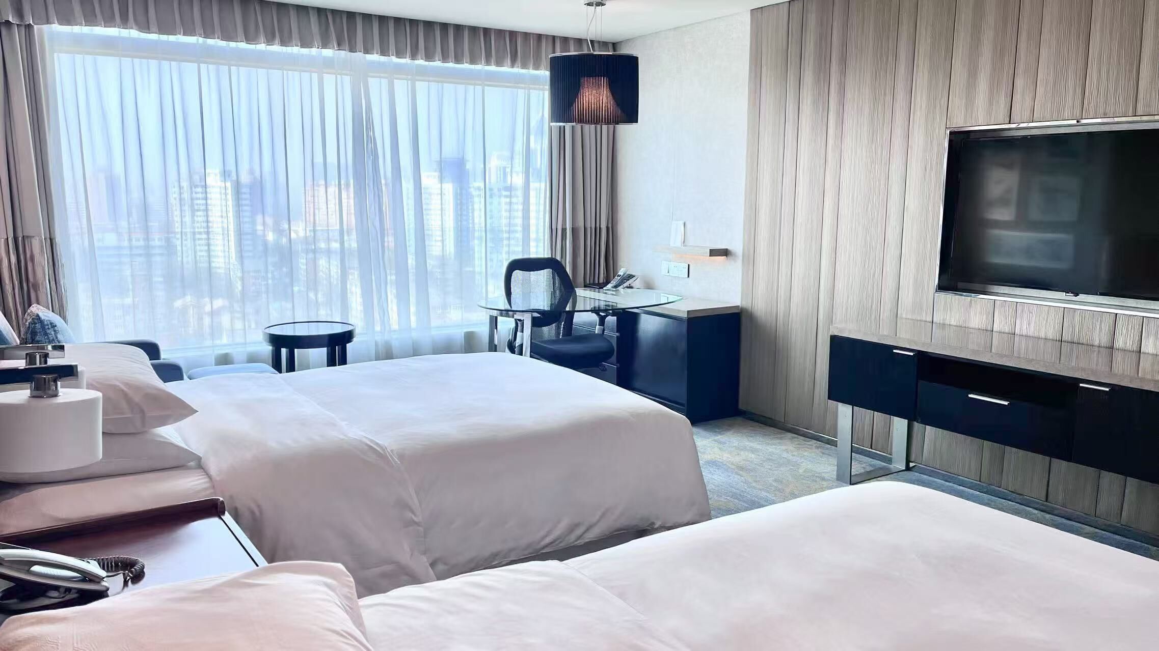 A hotel room with two beds, a desk, a chair, a TV, and a view of the city.