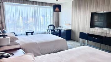 A hotel room with two beds, a desk, a chair, a TV, and a view of the city.
