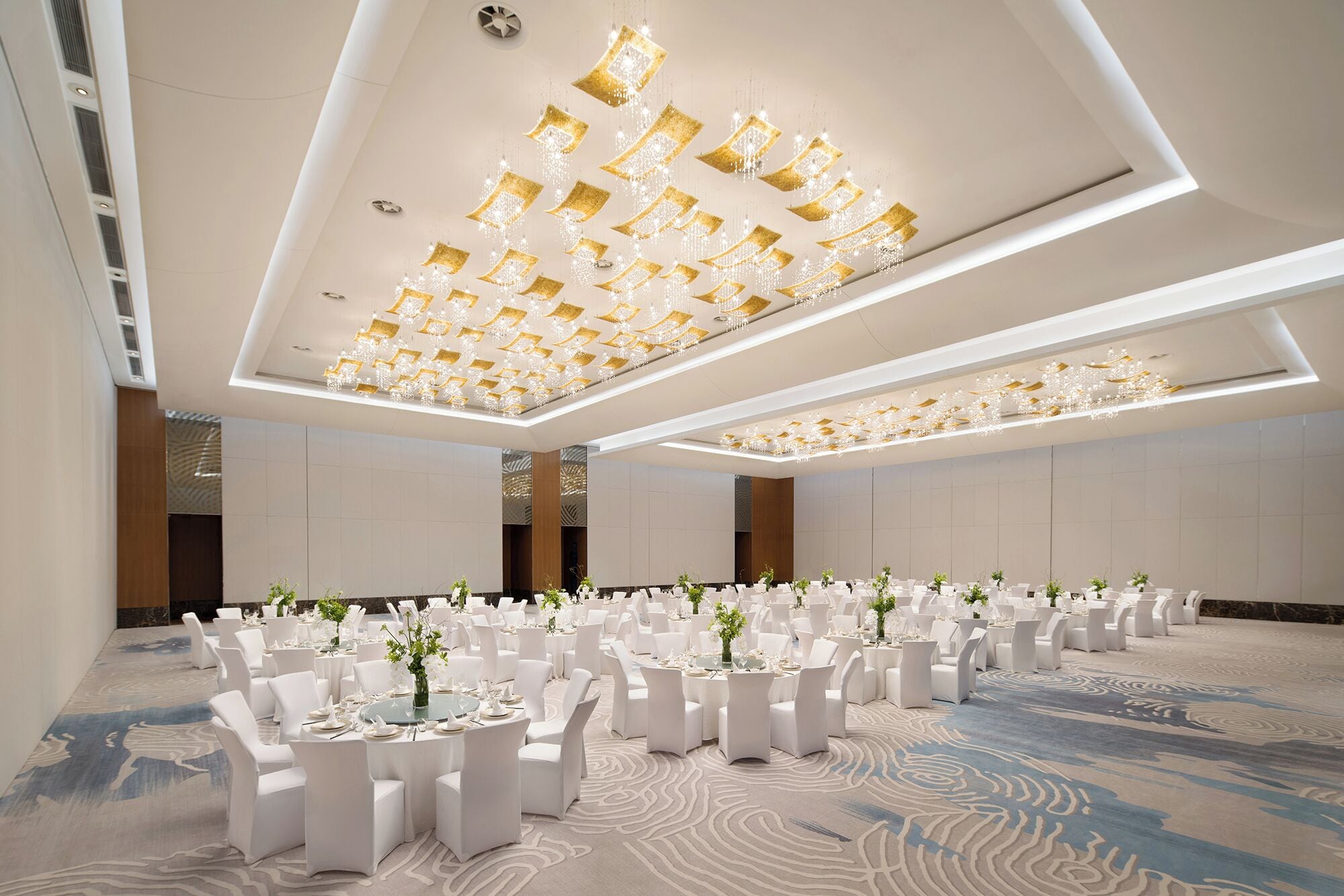 A banquet hall with round tables, white chairs, and a ceiling with geometric light fixtures.