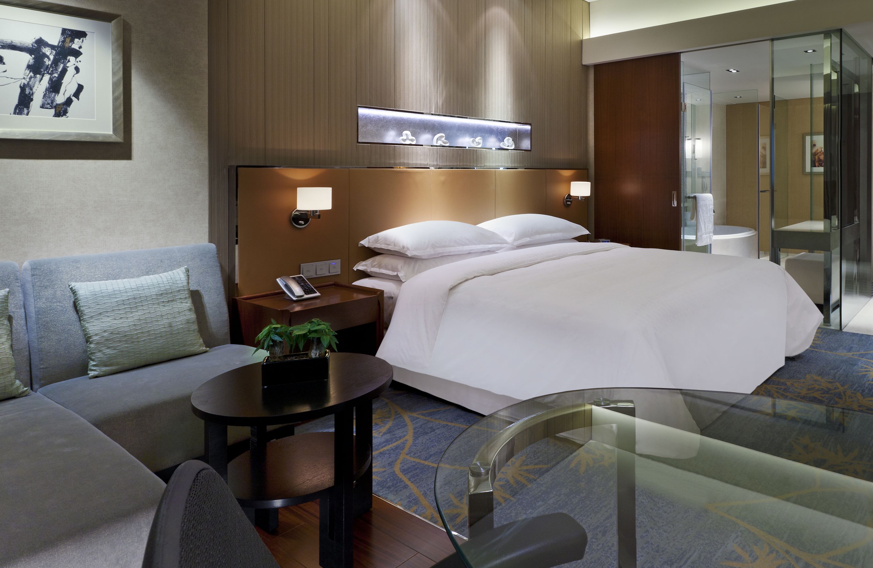 Una habitación de hotel moderna con una cama grande, un sofá, una mesa de vidrio y una mesita con una planta.