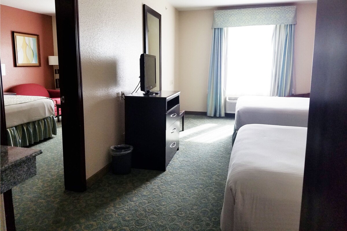 A hotel room with two beds, a desk with a television, a waste bin, and a window with curtains.