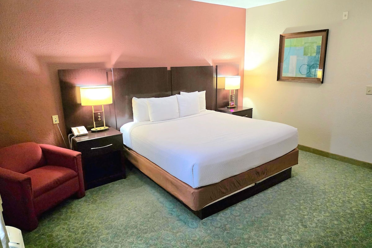 Un cuarto de hotel con una cama grande, dos lámparas de noche, un sillón rojo, una mesita con un teléfono y un cuadro en la pared.
