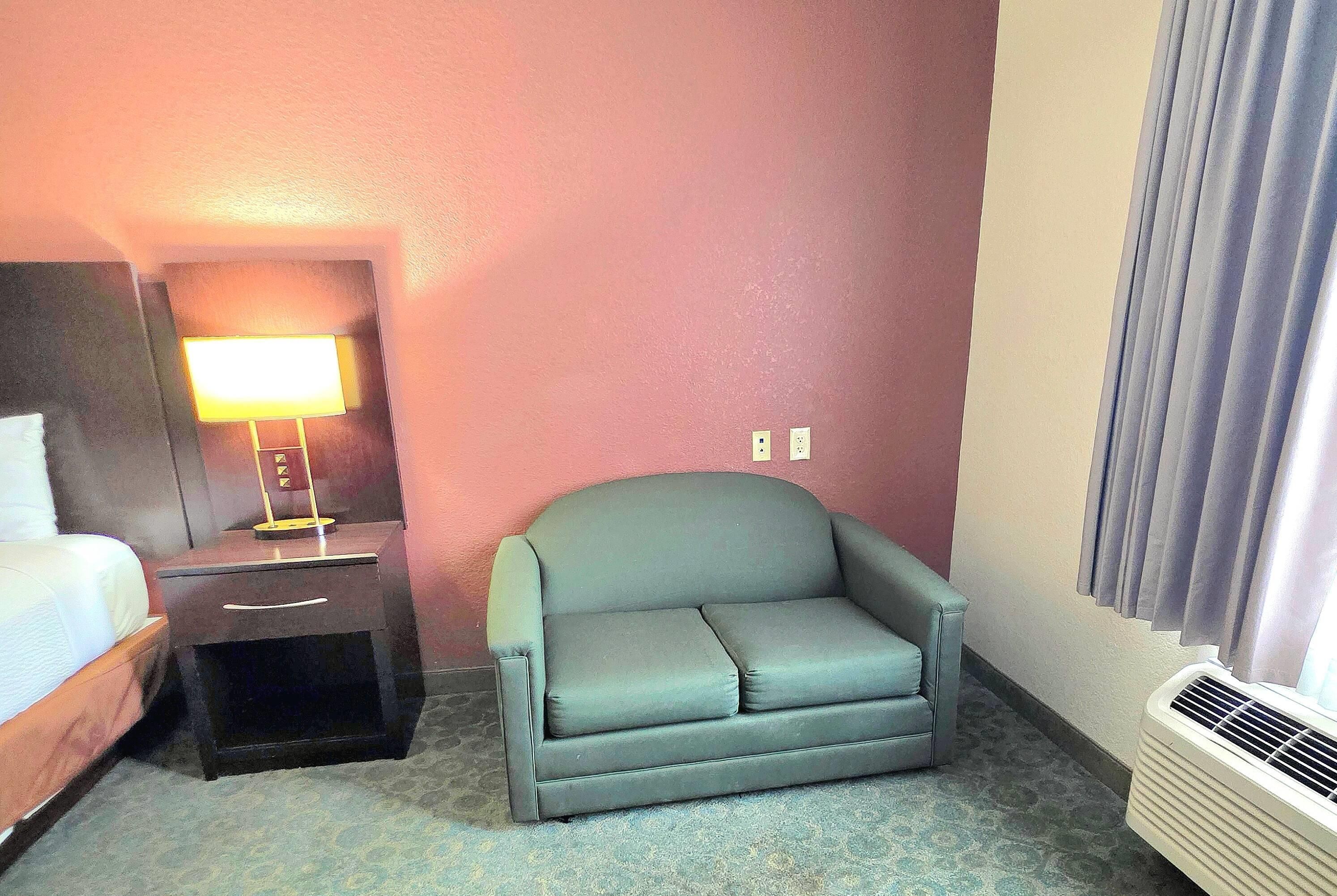 Una habitación de hotel con una cama, una mesita de noche con lámpara, un sillón gris y una ventana con cortinas.